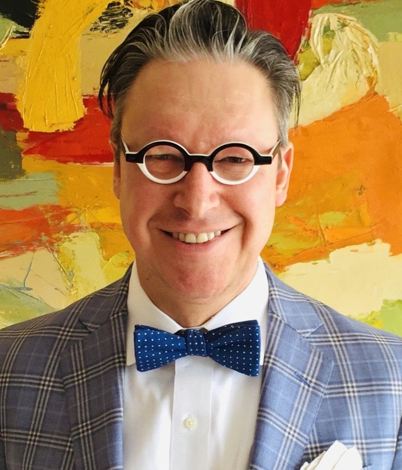 Trae Nunnink, smiling, in round glasses, a blue plaid jacket, and a navy polka-dot bow tie
